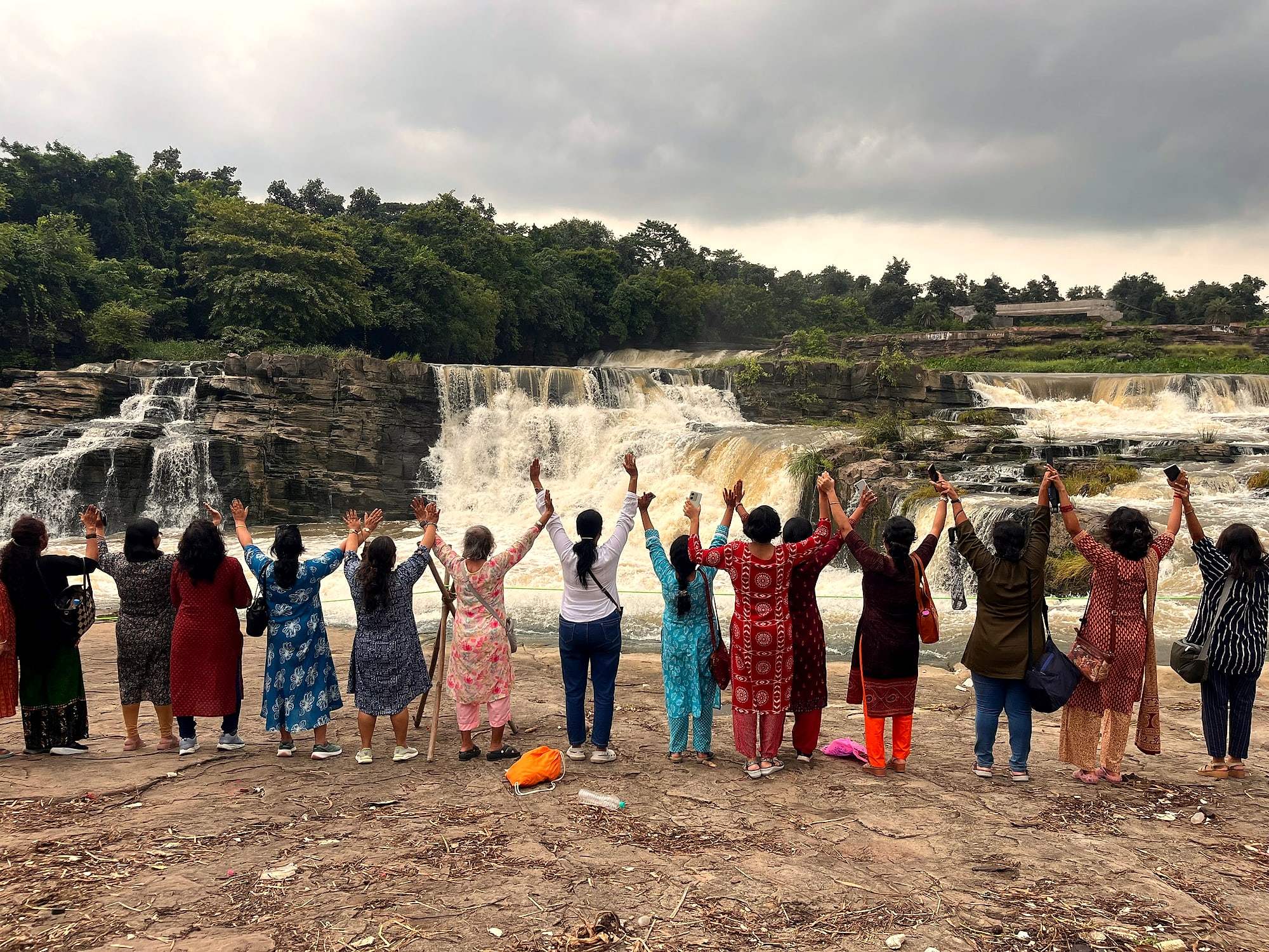 A group of ECHEPURON travellers cheering with arms raised in front of a waterfall